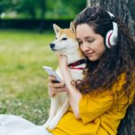 A woman in yellow cuddles a Shiba Inu while using a smartphone outdoors, enjoying music.