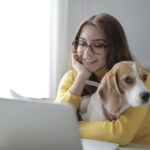 Happy woman in yellow sweater smiles while hugging her dog and using a laptop indoors.